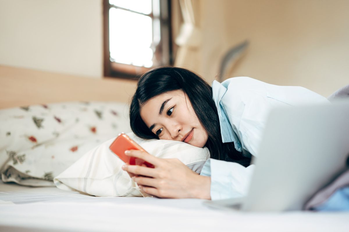 Seorang wanita sedang bermain ponsel sambil rebahan