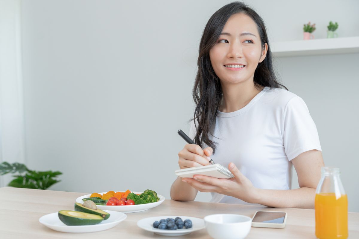 Wanita muda sedang duduk di meja makan sambil mencatat di notebook