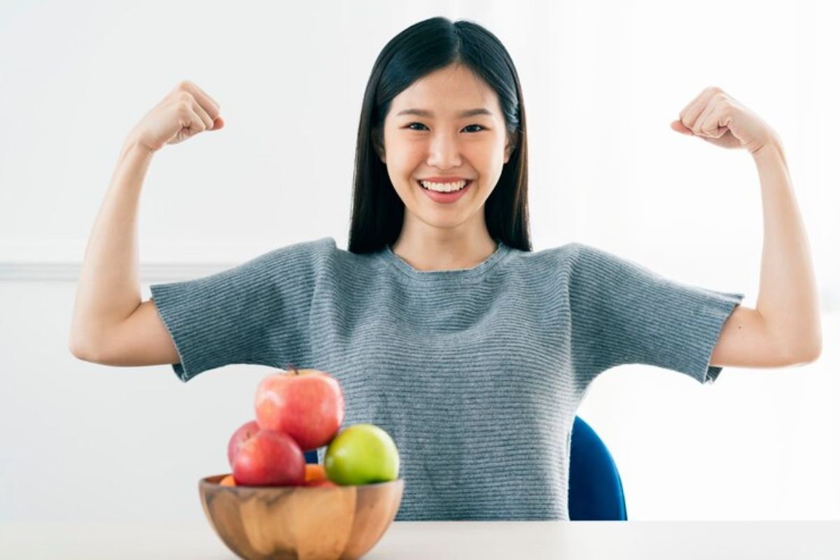 seorang wanita tersenyum sambil mengangkat tangan dan tumpukan buah apel di dalam keranjang di depannya