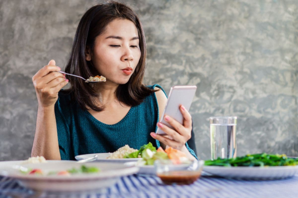 Wanita sedang makan sambil membawa ponsel