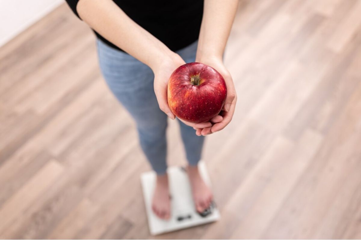 Perempuan berdiri di atas timbangan sambil memegang buah apel