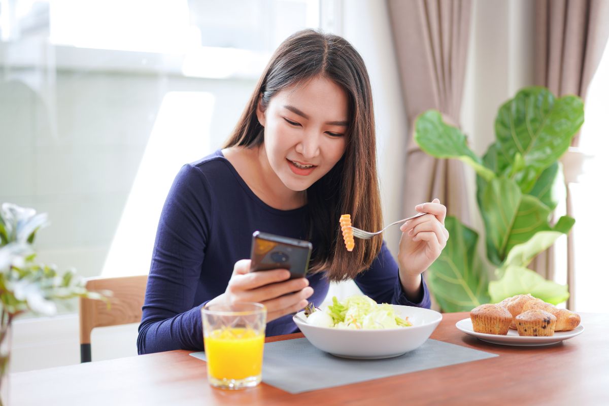 Seorang wanita sedang menikmati sarapan sambil bermain ponsel