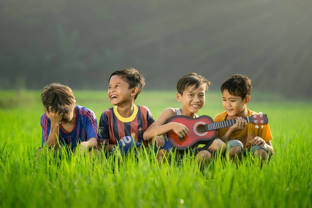 anak-anak yang sedang tertawa di tengah rumput sambil membawa gitar